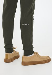 Beige suède schoenen met rubberen textuurzolen, gedragen met donkere groene joggers met witte tekst, waardoor een casual outfit ontstaat.