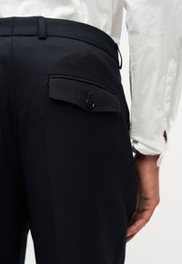 Κοντινό πλάνο ατόμου που φοράει σκούρο παντελόνι με κουμπωμένη πίσω τσέπη και λευκό πουκάμισο με μακριά μανίκια, με ορατά τα κουμπιά στις μανσέτες.