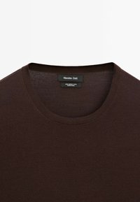 Maglione in lana merino marrone con scollatura rotonda. Presenta una texture a coste attorno al colletto e all'orlo, e un design minimalista.