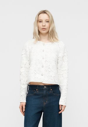 Blonde Frau trägt eine weiße, mit Blumenmuster verzierte Spitzenbluse mit Knöpfen und hoch taillierte dunkelblaue Jeans, steht vor einem einfarbigen Hintergrund.