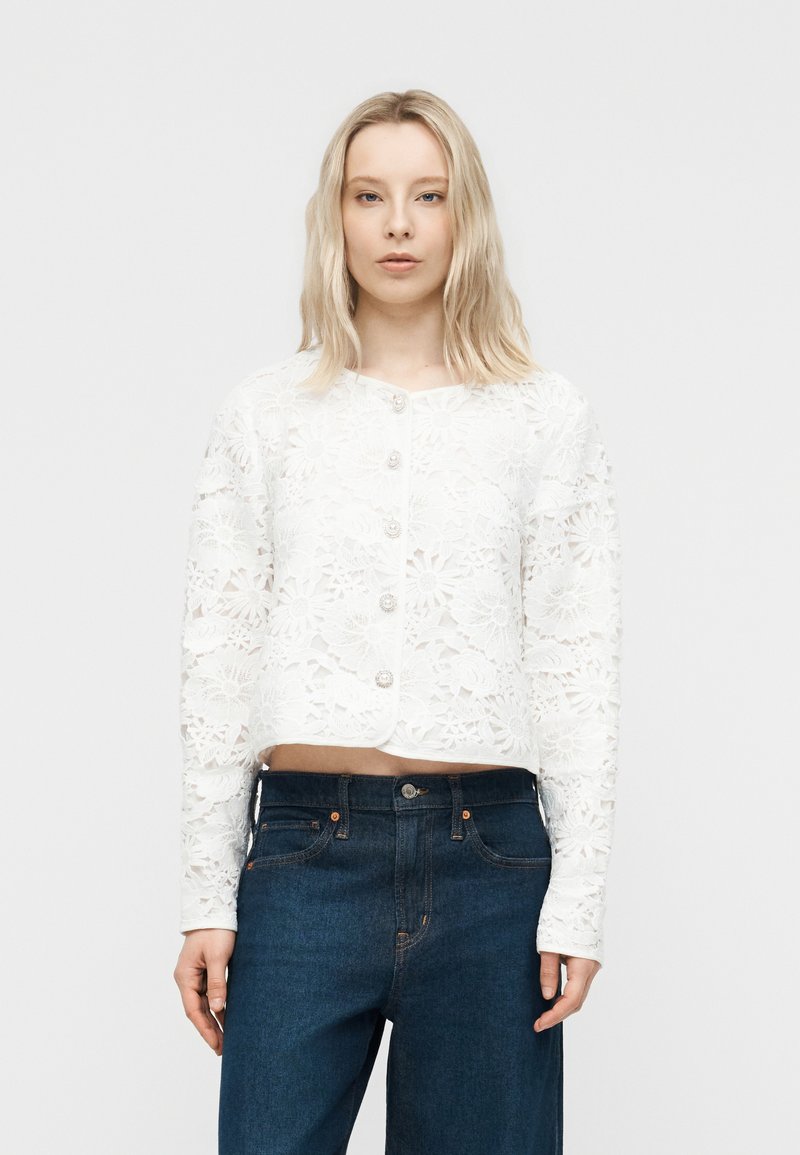 Heledate juustega naine, kes kannab valget lillelise pitsiga nööbitavat pluusi ja kõrge vöökohaga tumelisi siniseid teksapükse, seisab ühevärvilise tausta ees.