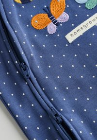 Blå bomullsjacka med dragkedja och vita prickar, med broderade fjärilar i orange och lila, samt en tagg med texten "hemodlad".