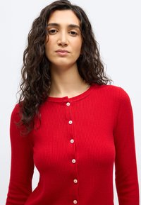 Νεαρή γυναίκα με μακριά σγουρά σκούρα μαλλιά, φορώντας κόκκινο ριπ πουλόβερ με κουμπιά, πάνω σε απλό φωτεινό φόντο.