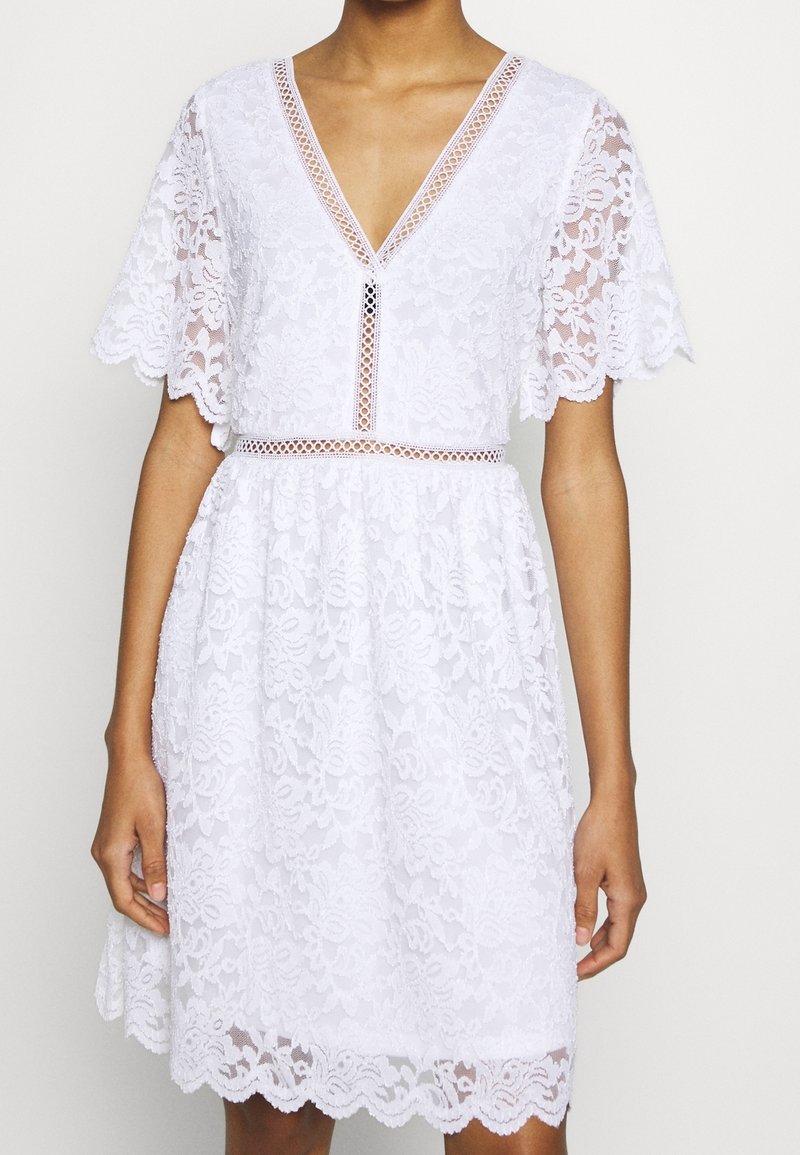 Robe blanche en dentelle avec manches volantées, col en V et ourlet festonné, portée par une personne devant un fond clair uni.