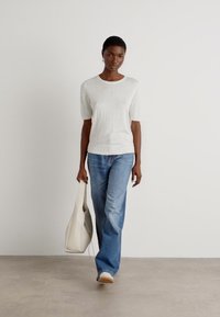 Un haut en tricot gris clair à manches courtes associé à un jean bleu moyen à jambe droite. Le mannequin porte un sac fourre-tout crème doux et des baskets blanches.