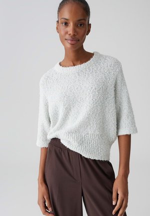 Vrouw die een witte, gestructureerde trui met korte mouwen en een donkerbruine, high-waisted broek draagt tegen een effen grijze achtergrond.