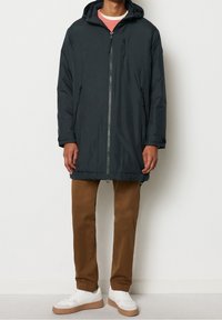 Giubbotto piumino blu scuro con cappuccio, zip anteriore e tasche laterali, abbinato a pantaloni marroni e sneakers bianche. Texture liscia.