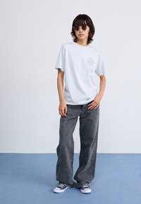 Lyseblå bomulls T-skjorte med trykk på venstre bryst, kombinert med løse grå denimjeans og marineblå sneakers. Runde svarte solbriller på.