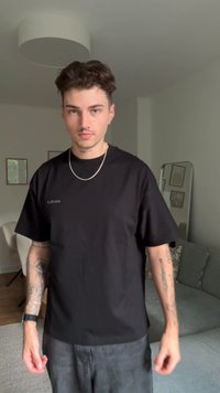 T-shirt preto de manga curta com um corte solto e detalhe de logo discreto na frente. O modelo tem tatuagens e está a usar joias de corrente de prata.