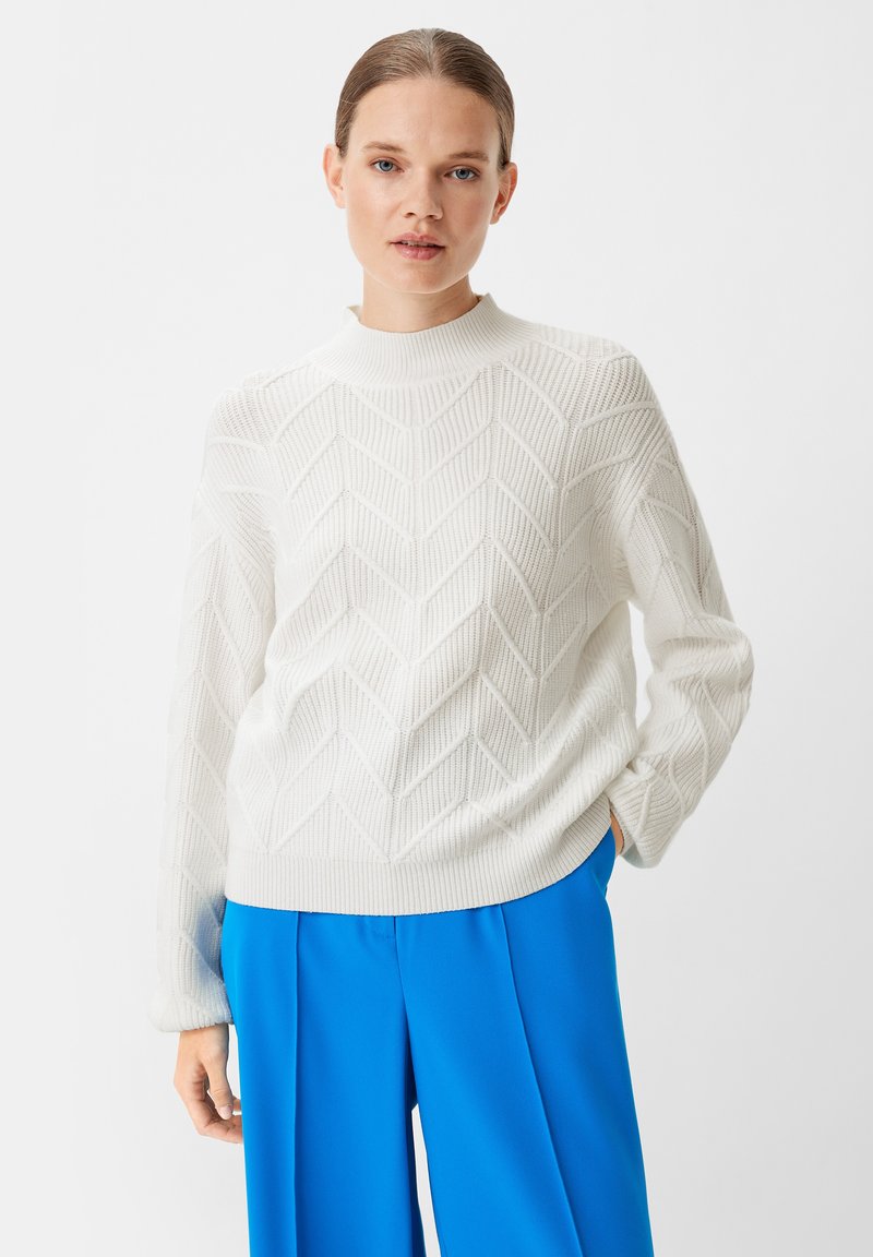 Weißer Strickpullover mit einem strukturierten Zickzackmuster und hohem Kragen, kombiniert mit leuchtend blauen weitem Hosen.