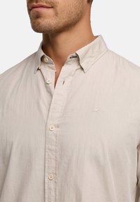 Beige Hemd mit Knöpfen, klassischem Kragen, leichtem Stoff, feiner Textur und kleinem gesticktem Logo auf der Brust.