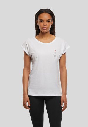 Femme portant un t-shirt blanc à manches retroussées avec un petit motif main-cœur sur la poitrine gauche, associée à des leggings noirs, debout devant un fond gris.