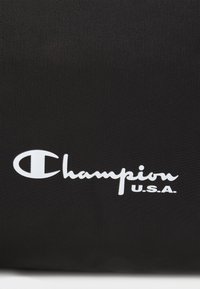 Črna najlonska torba z belim vezenim logotipom "Champion" in besedilom "U.S.A.", poudarjena z gladko teksturo in športnim dizajnom.