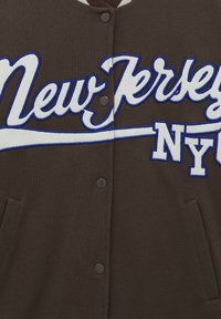 Brązowa bluza z kapturem z białym i niebieskim haftowanym napisem "New Jersey NY." Posiada napy i boczne kieszenie. Miękka tekstura materiału.
