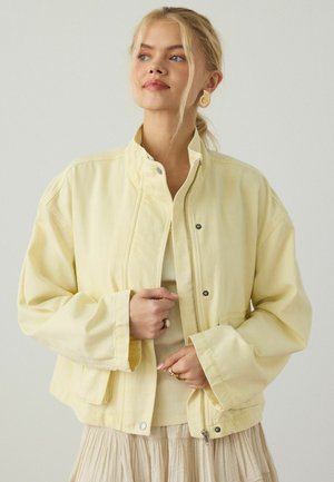 Blondynka w jasnop żółtej kurtce na beżowy top i spódnicę, poprawia suwak kurtki na jednolitym jasnym tle.