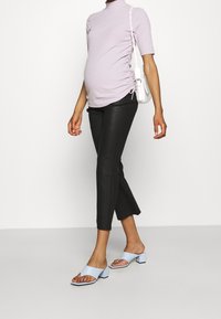 Donna incinta che indossa un top lilla arricciato, pantaloni neri a vita alta, sandali con tacco azzurri chiari e porta una borsa a spalla bianca.