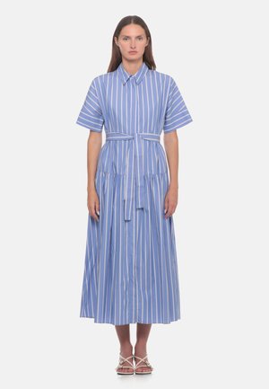 Donna in piedi che indossa un vestito midi a righe verticali blu e bianche con maniche corte, colletto, cintura in vita e sandali bianchi open toe.