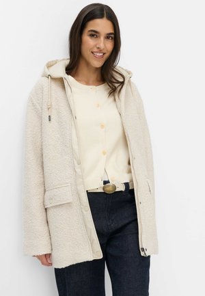 Frau lächelt, trägt einen cremefarbenen, strukturierten Kapuzenmantel über einem cremefarbenen Cardigan und dunkle Jeans mit Gürtel vor einem einfarbigen weißen Hintergrund.