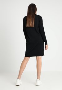 Svartstickad klänning med långa ärmar och ribbade detaljer på axlarna. Kjolens nederkant är rak och slutar strax ovanför knäna, i kombination med vita skor.