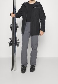 Czarna wodoodporna kurtka, szare spodnie z łatami na kolanach, trzymające narty. Buty narciarskie o czarnym, błyszczącym wykończeniu i regulowanymi klamrami. Gładkie tekstury.