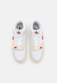 Witte sneaker van geperforeerd leer, met een rode Nike-logo, beige strap en zool, en contrasterende zwarte accenten op de binnenvoering.