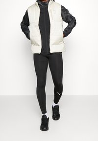 Padded kremowa kamizelka z kapturem na czarnej kurtce zapinanej na zamek. Czarne dopasowane legginsy i czarne buty sportowe z białymi podeszwami.