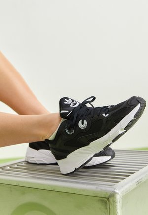 Piernas con zapatillas Adidas negras y blancas con malla y cordones, descansando sobre una superficie metálica con un fondo liso.