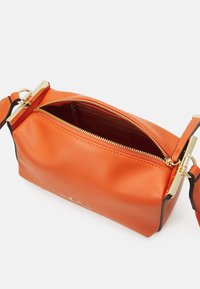 Sac en cuir orange avec fermeture éclair et éléments en or. Texture lisse, forme structurée et détail du logo sur le côté. Sangle d'accessoire incluse.