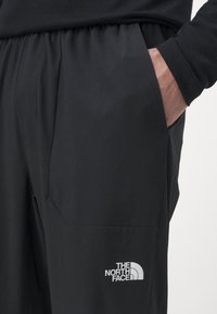 Fekete nadrág könnyű anyagból, oldalsó zsebbel és jól látható fehér The North Face logóval a lábszáron.