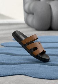 Sandale marron synthétique à glissière avec double bride et design ajouré, semelle en caoutchouc noir, posée sur un tapis texturé bleu.