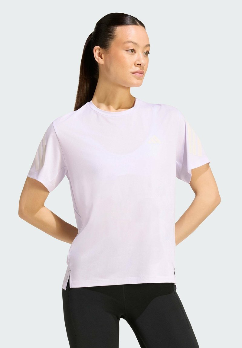 Femme aux longs cheveux foncés attachés en queue de cheval, portant un t-shirt violet clair à manches courtes et des leggings noirs, debout les mains sur les hanches, regardant vers la droite.