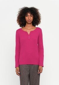 Magenta langærmet top med Henley-halsudskæring og tre knapper. Har en ribbet struktur og er parret med grå mønstrede bukser.