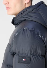 Giacca puffa blu con cappuccio, caratterizzata da una texture lucida e un design trapuntato. Patch del logo Tommy Hilfiger sul braccio.