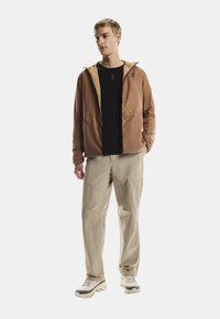 Joven con una chaqueta marrón con cremallera, camiseta negra, pantalones beiges y zapatillas blancas, de pie frente a un fondo blanco liso.
