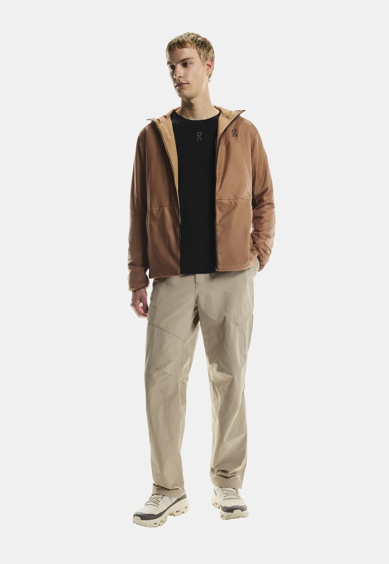 Joven con una chaqueta marrón con cremallera, camiseta negra, pantalones beiges y zapatillas blancas, de pie frente a un fondo blanco liso.