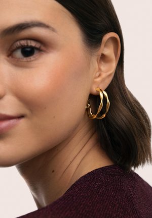 Mujer con cabello castaño corto que lleva pendientes de aro dorados retorcidos y una blusa texturizada de color púrpura oscuro, mostrada en un primer plano de perfil.