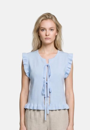 Vrouw draagt een lichtblauw mouwloos topje met geruite randen en twee strikjes aan de voorkant, gecombineerd met beige broek, staand tegen een witte achtergrond.