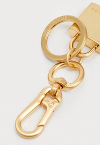 Gold metal keychain with a flat keyring, oval connector, and a hook clasp. Features embossed brand name and smooth metallic texture.