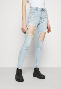 Ljusblå skinny jeans med slitna knän och fransade kanter. Kombineras med svarta ankelstövlar, visad mot en neutral bakgrund.