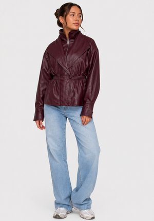 Kvinde står iført en mørkerød jakke i kunstlæder med lynlås samt blå vidde jeans og hvide sneakers mod en ensfarvet baggrund.