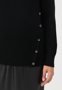 Black long-sleeve sweater with side button closure detail worn over a black leather skirt, featuring a visible hand on the right side.