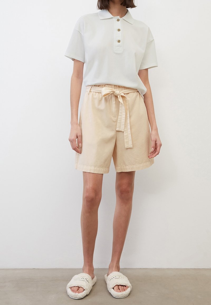Femme portant un polo blanc à manches courtes, un short beige taille haute avec une ceinture nouée, et des chaussons blancs pelucheux à bout ouvert.