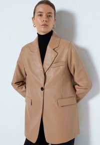 Beige læder blazer med en struktureret pasform, enkelt sort knaplukning, to forlommer og klippede revers. Bæres over en sort rullekrave.