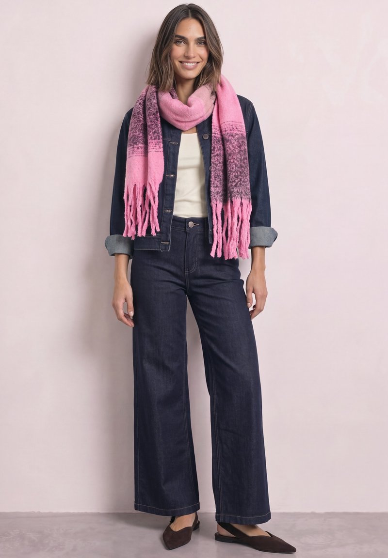 Femme souriante portant une veste en jean foncée et un jean à jambes larges avec un haut blanc, un foulard rose à motifs et des chaussures pointues marron sur fond uni.