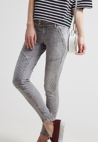 Jeans skinny de mezclilla gris con parches texturizados en los muslos, un ligero desgaste y una silueta ajustada. Combinados con un bolso cruzado blanco.