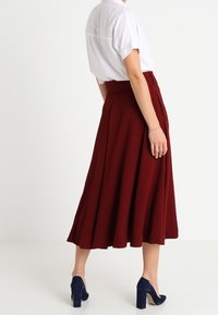 Jupe A-line bordeaux avec des plis, en tissu fluide, associée à une chemise blanche à manches courtes et des chaussures à talons hauts bleues.