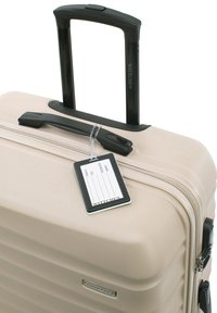 Maleta rígida beige con asa retráctil negra, asa superior, cremallera y etiqueta de plástico para equipaje adjunta con campos para nombre y contacto.