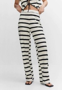 Femme portant un pantalon en tricot rayé noir et blanc à rayures horizontales et un haut sans manches assorti, se tenant avec les mains ajustant la taille.