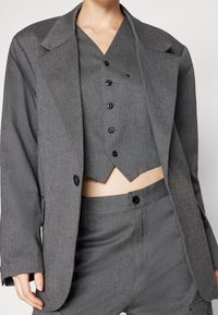 Szary teksturowany garnitur zawierający dopasowaną marynarkę, skrócony kamizelkę z ostrym dołem oraz dopasowane spodnie z czarnymi guzikami i akcentami.