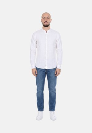 Uomo in piedi di fronte a noi, che indossa una camicia bianca abbottonata, jeans blu e scarpe da ginnastica bianche su uno sfondo chiaro e semplice.